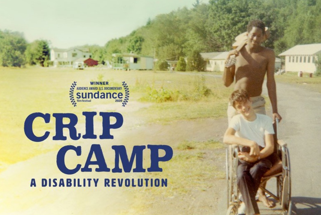 Image of Crip Camp movie poster. A young man sits in a wheelchair while another young man, who is holding a guitar over his shoulder, stands behind him. 